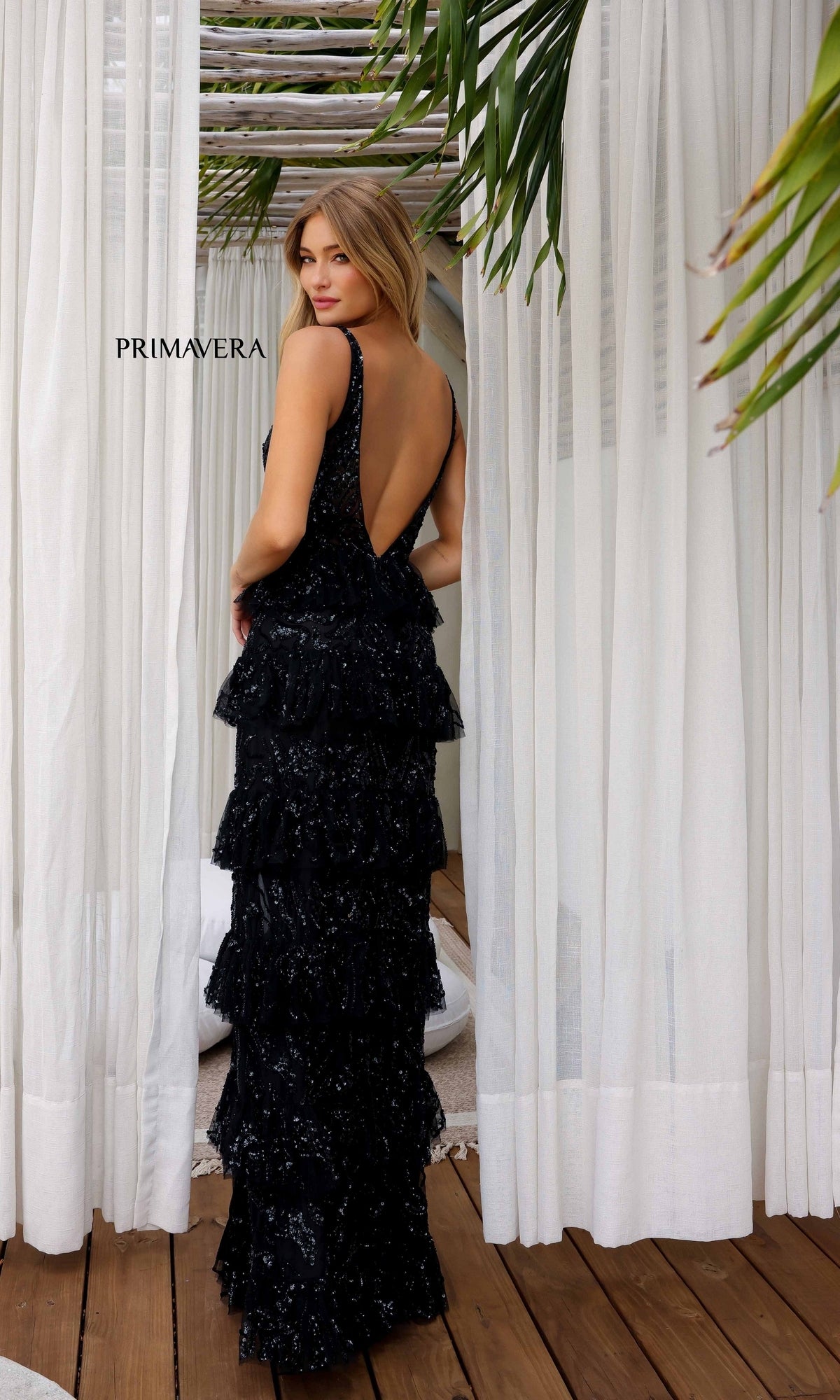 A woman stands between sheer white curtains and green palm leaves, wearing the Long Formal Dress 4295 by Primavera—a black, ruffle-layered, backless sequin gown.